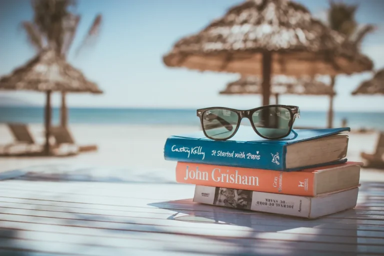 A stack of books with sunglasses on top sits on a white table by the beach, with palm trees and umbrellas in the background, evoking a relaxing vacation mood