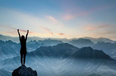 A silhouette of a person standing on a mountain peak with arms raised in victory, overlooking a scenic view of misty mountains at sunrise.