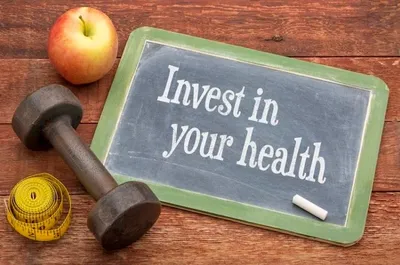 "Chalkboard with 'Invest in your health' written on it, placed on a wooden surface alongside a dumbbell, an apple, and a measuring tape."